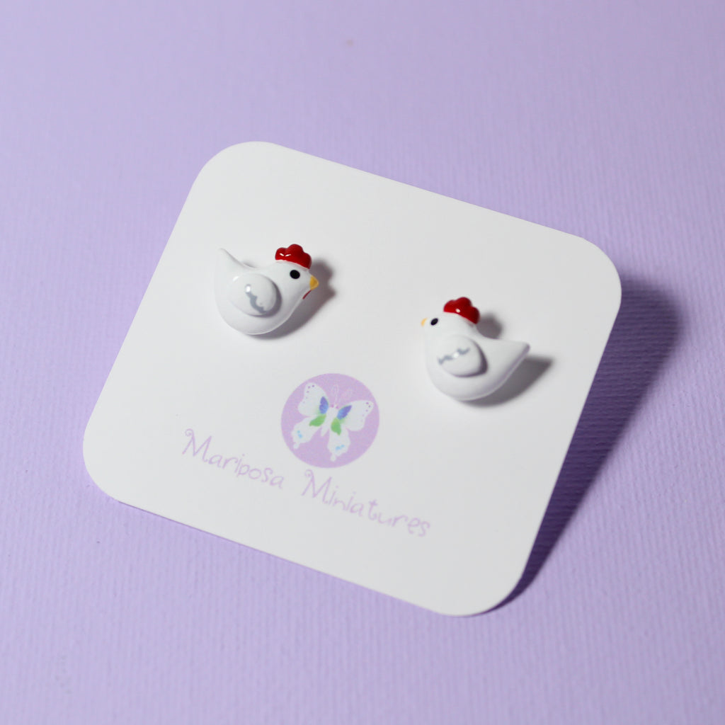 A pair of chicken stud earrings are displayed on a white jewelry backing card