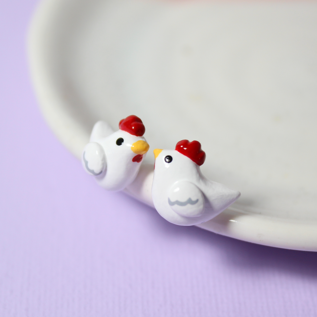 A pair of white chicken stud earrings rest against a white jewelry dish