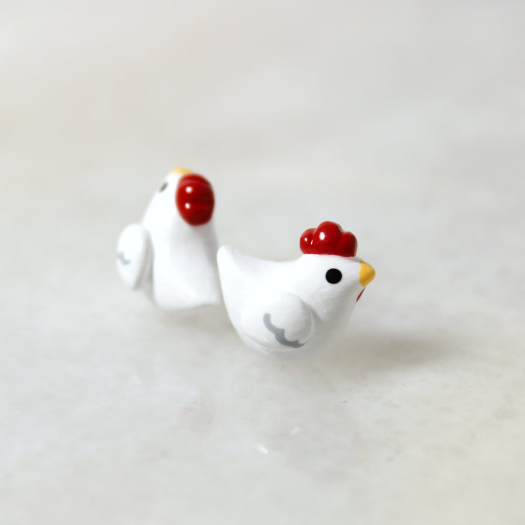 A pair of chicken stud earrings rest on top of a marble surface