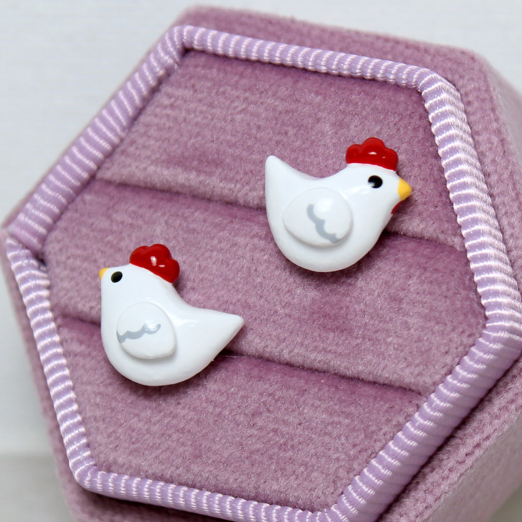 A pair of chicken stud earrings are displayed in a hexagonal purple velvet jewelry box