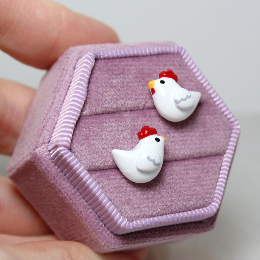 A hand holds a hexagonal purple velvet jewelry box. A pair of chicken stud earrings are nestled in the jewelry box