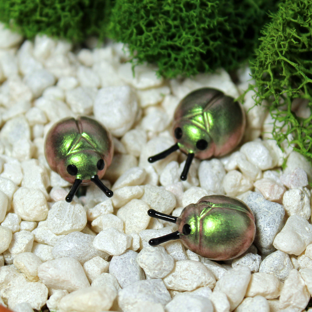 Three miniature beetle figurines sit in a moss garden. The beetles have iridescent green/gold shells