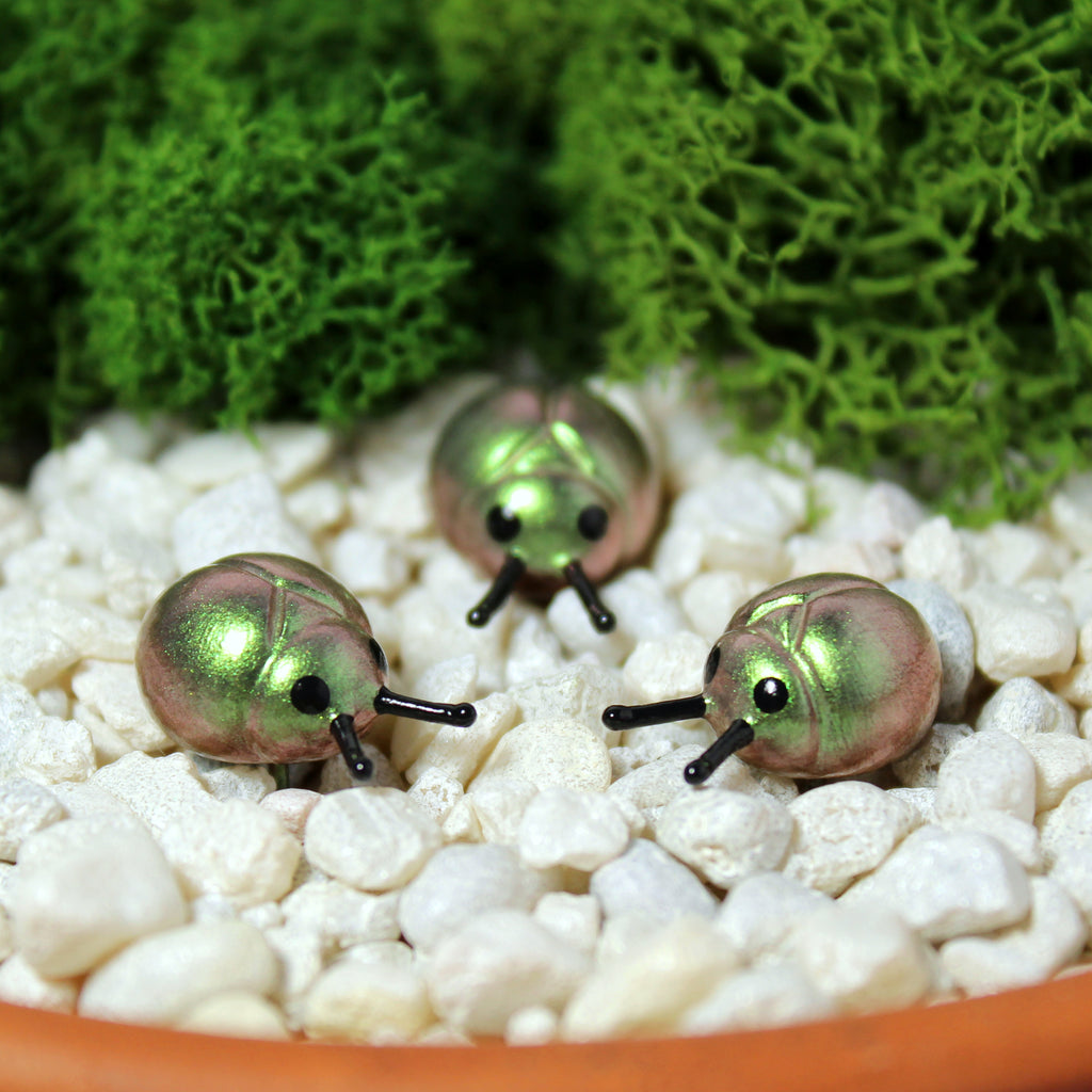 Three miniature beetle figurines sit in a moss garden. The beetles have iridescent green/gold shells