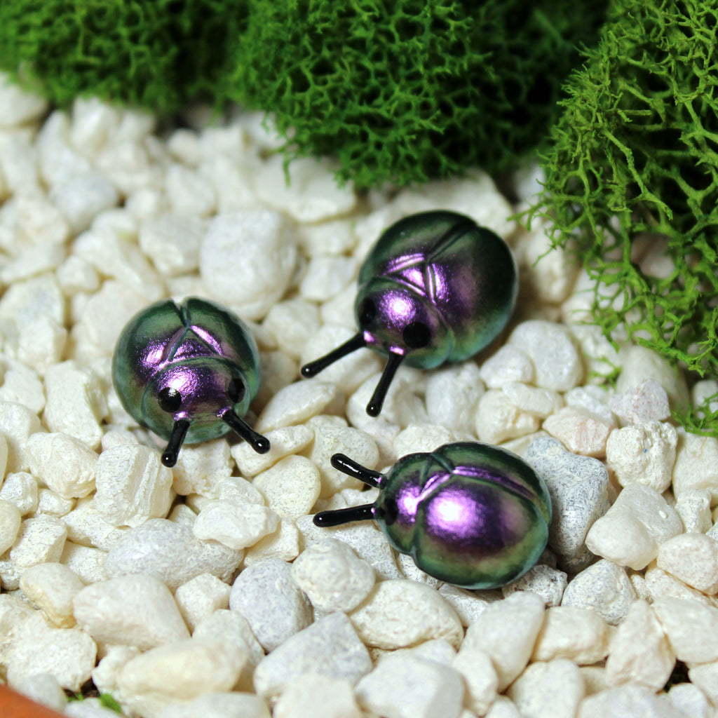 Three miniature beetle figurines sit in a moss garden. The beetles have iridescent purple/silver shells