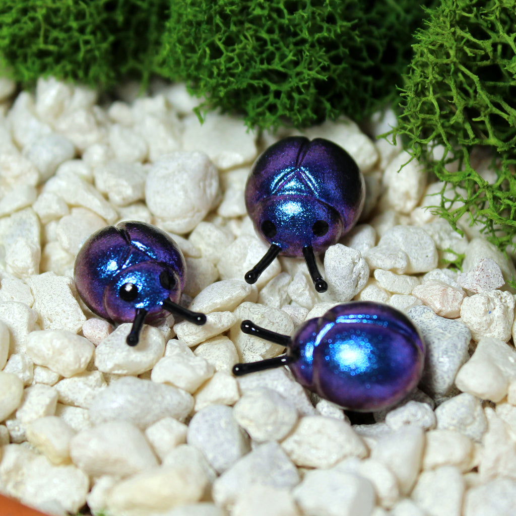 Three miniature beetle figurines sit in a moss garden. The beetles have iridescent blue/purple shells