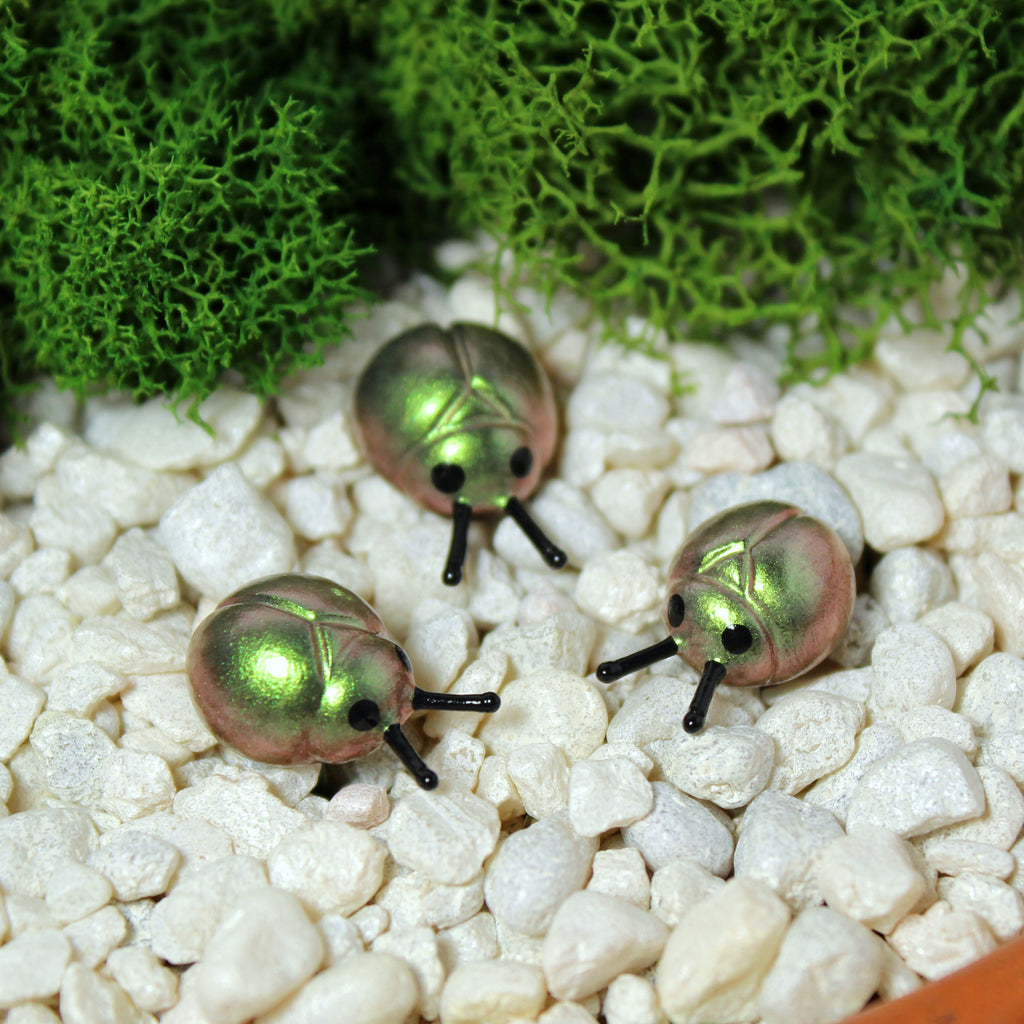 Three miniature beetle figurines sit in a moss garden. The beetles have iridescent green/gold shells