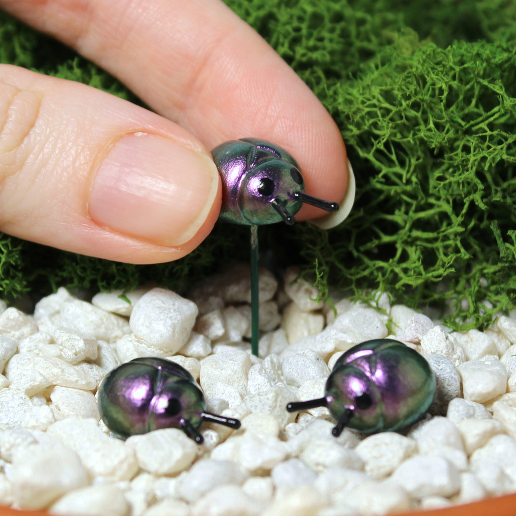 A hand places a miniature beetle figurine in a moss garden. The beetles are about half an inch wide with inch long wire stakes