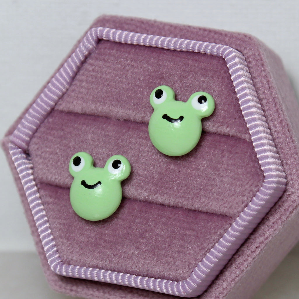 A pair of green frog stud earrings are displayed in a hexagonal purple velvet jewelry box