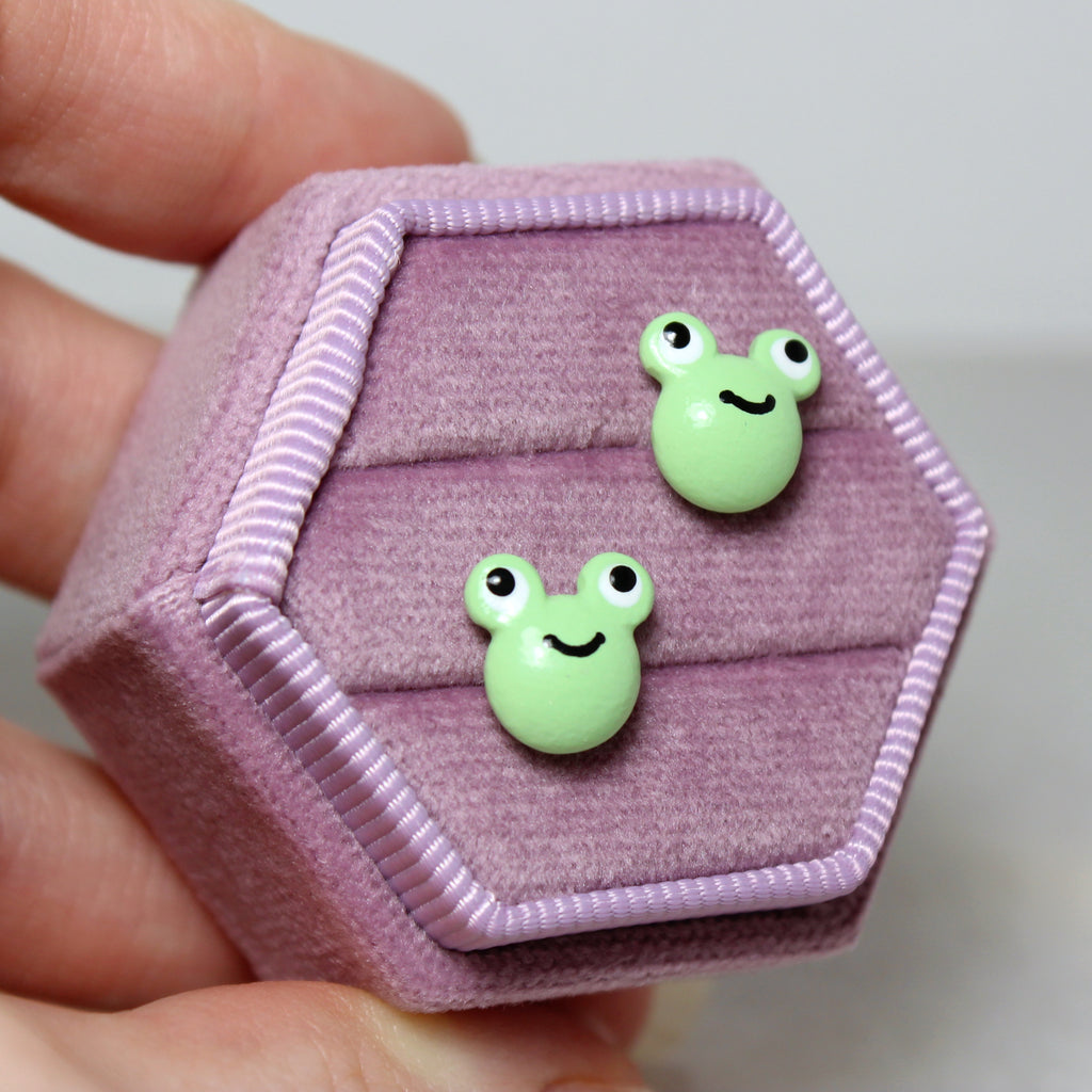 A hand holds a hexagonal purple velvet jewelry box. A pair of green frog stud earrings are displayed in the box