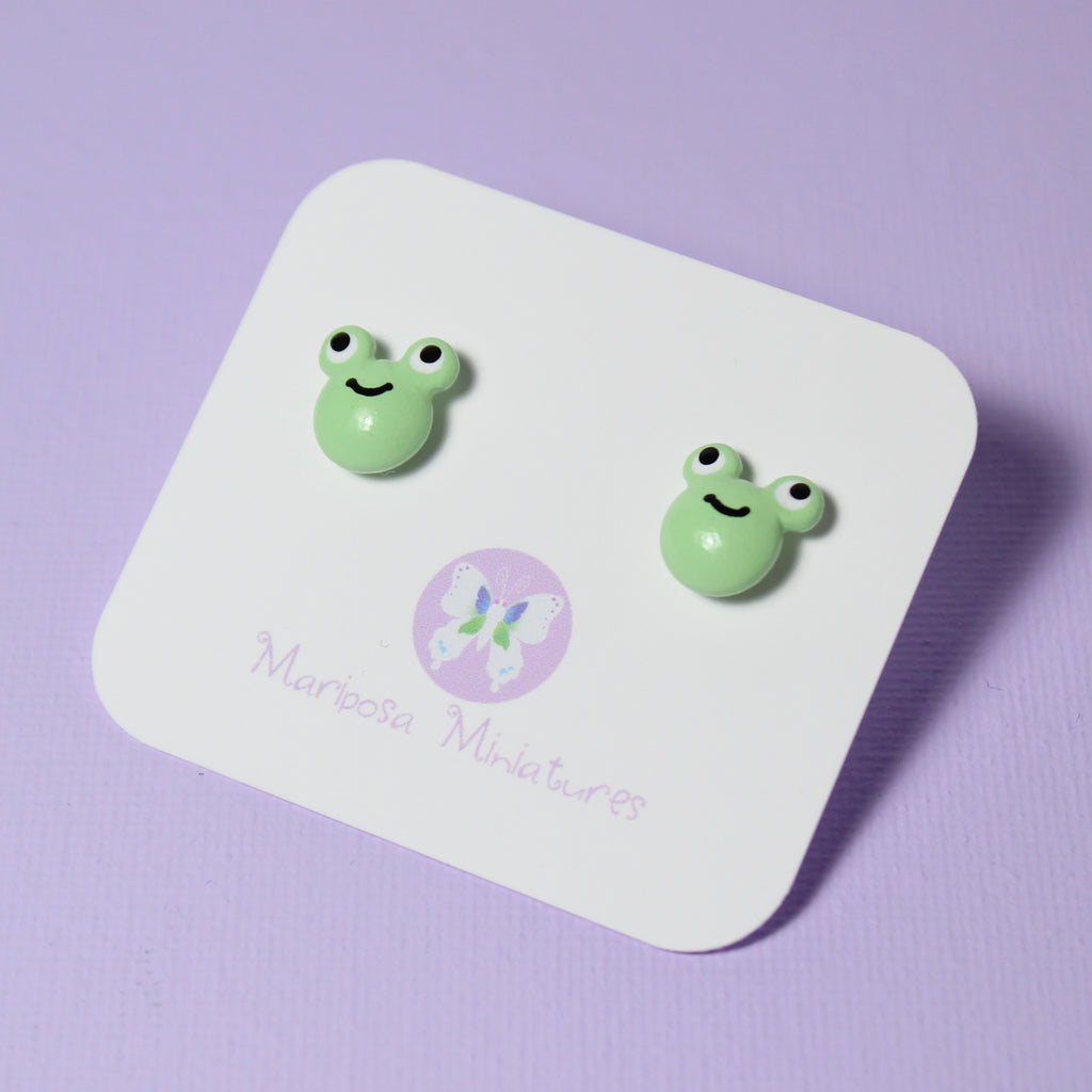 A pair of frog stud earrings are attached to a jewelry backing card