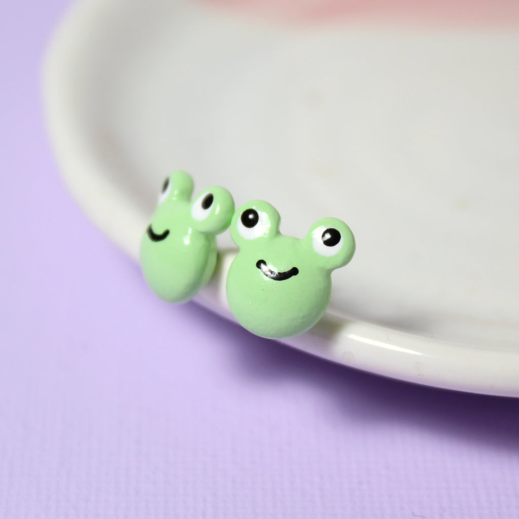 A pair of smiling green frog stud earrings rest against a white jewelry dish