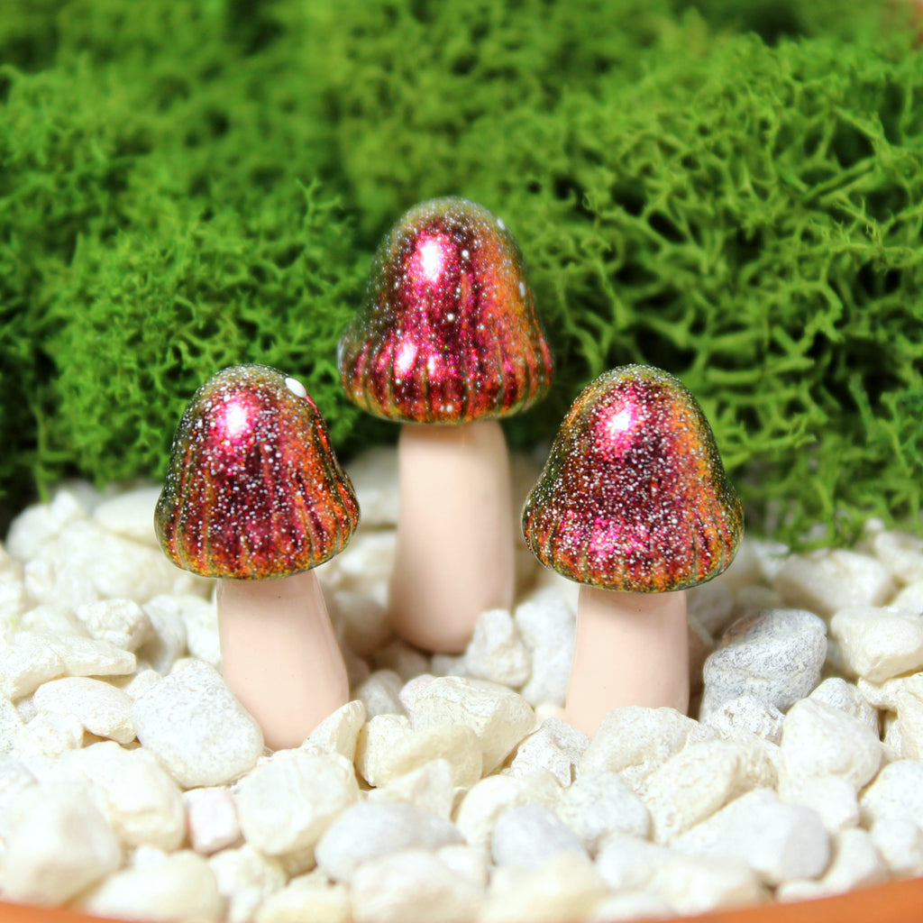 Three iridescent red and gold fairy mushrooms sit in a moss garden with white gravel